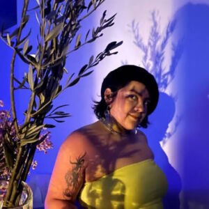 gg, lighting designer/artist mestiza tan non binary person with short dark hair and black beret hat, neon green sleeveless top standing in the shadow of some dried herb branches. the branches are seen to the left. the surrounding wall color is deep blue.