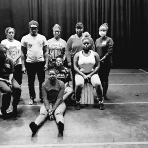 Gbedu Town Radio Ensemble in rehearsals at Counterpulse, San Francisco practicing a family portrait scene for the performance piece. Front row (R-L): Ebonie sitting on a white crate, Kanukai sitting on a stool slightly behind Uzo who is sitting in the front center, and Danaite sitting on the far left on a white crate. Back row (R-L): All standing, Piwai, KaliMa, Jameelah, Carmu, Roshonda, and Nkan Music.