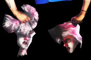 A white woman with bright makeup, pink hair, and a blue outfit on a black background is made into a lifesize paper doll holding her own head in both of her hands.