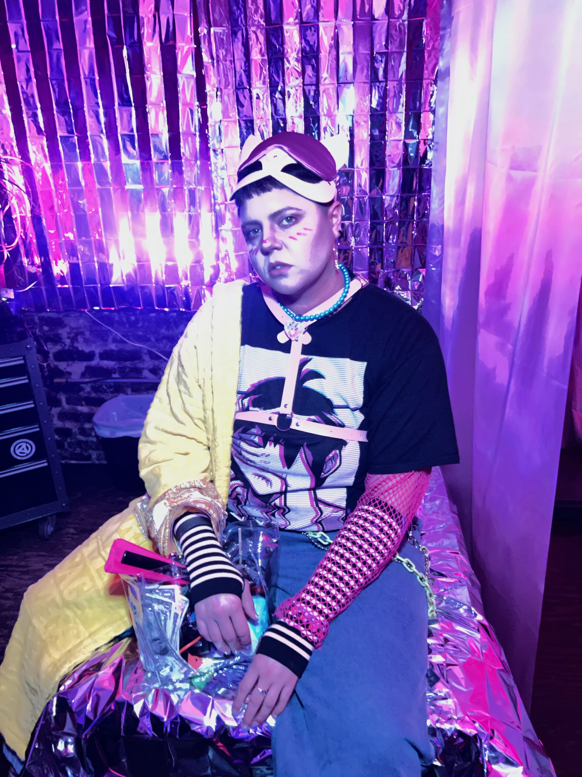 Alt Text: A white man white short hair sitting on top of a bed covered in tin foil and surrounded by silver streamers with a pink cat mask in colorful raver clothing. Alt Text: A white man white short hair sitting on top of a bed covered in tin foil and surrounded by silver streamers with a pink cat mask in colorful raver clothing.