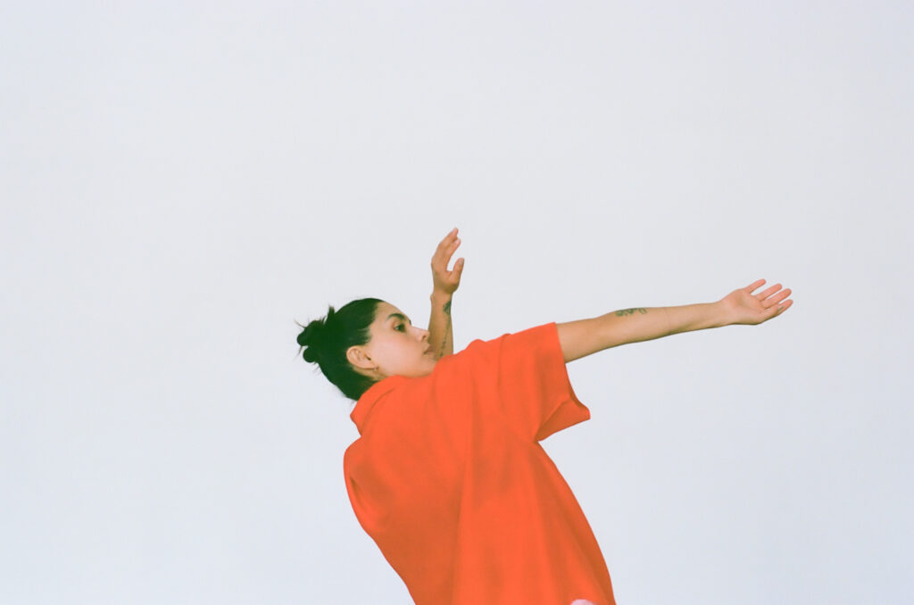 Photo of a person in an orange collared shirt, with black hair tied back, gesturing with hands above their head on a white background.