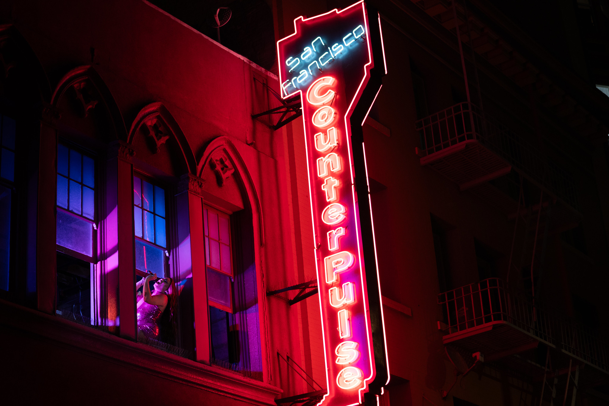 A drag performer sings through the windows of CounterPulse, with it's neon lights illuminating the building