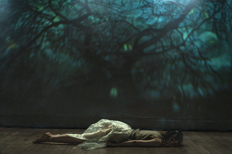 A dancer in a filmy light green costume lies flat, face down, on stage with a projection of a night forest over her.